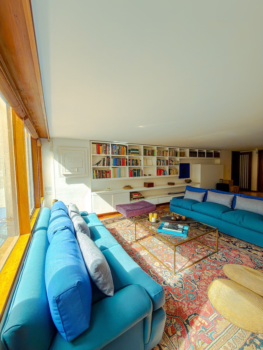 Sala junto al ventanal, con sofás azules enfrentados, alfombra estampada, mesa central y biblioteca blanca al fondo.
