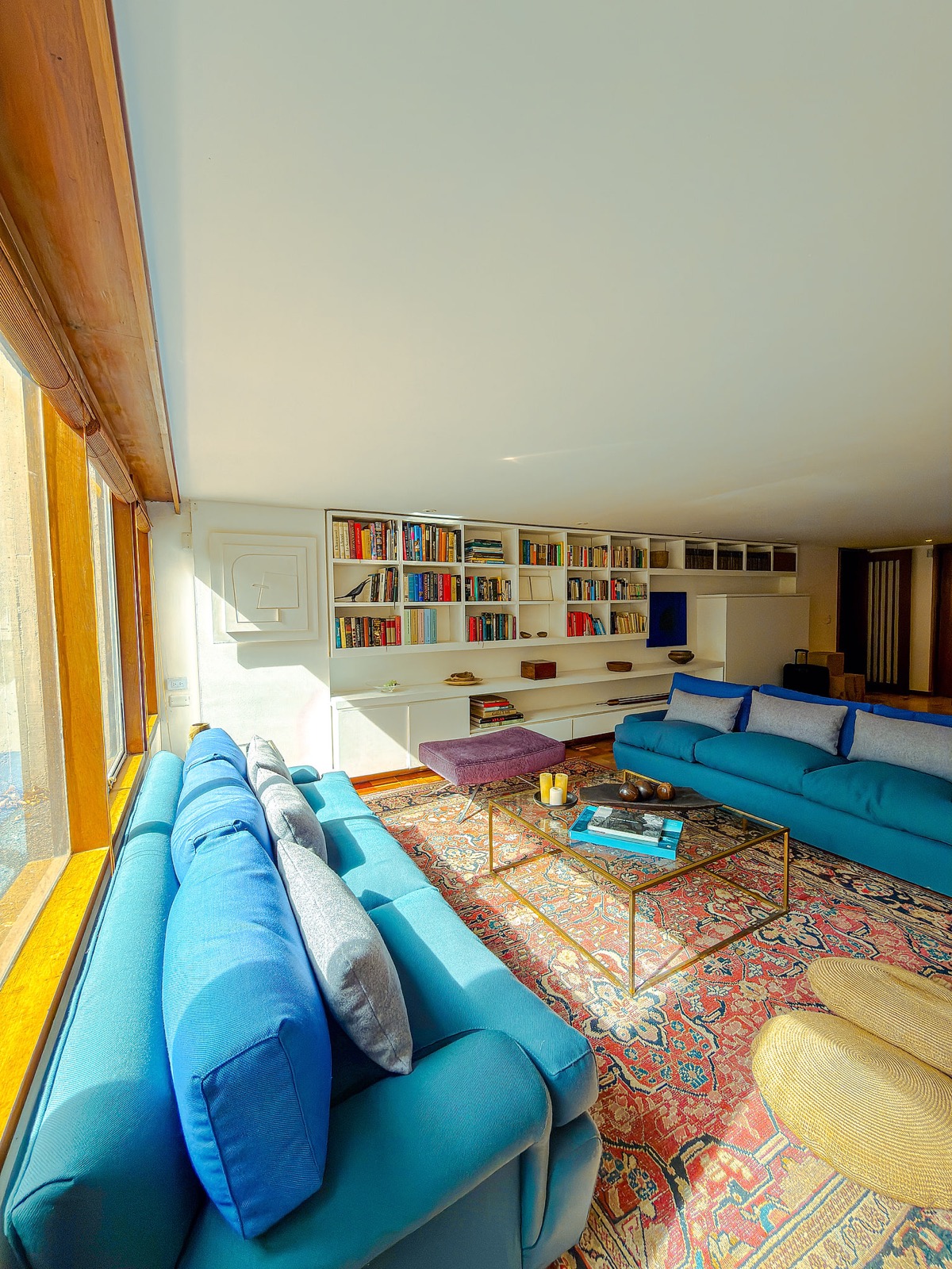 Sala junto al ventanal, con sofás azules enfrentados, alfombra estampada, mesa central y biblioteca blanca al fondo.