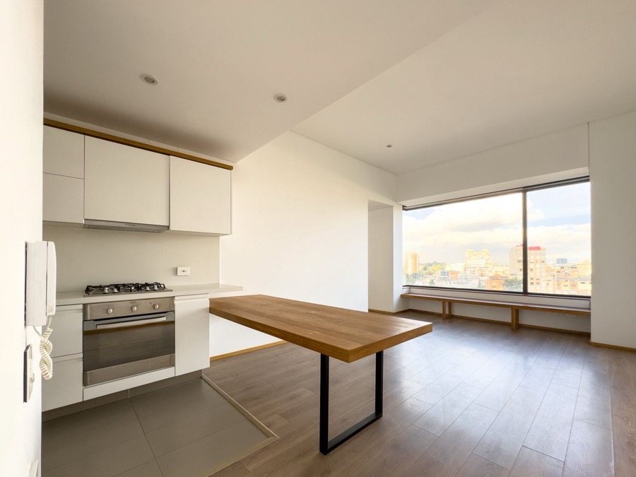 Vista general de cocina abierta y zona social, con mesón de madera, ventanal amplio y banca junto a la ventana.